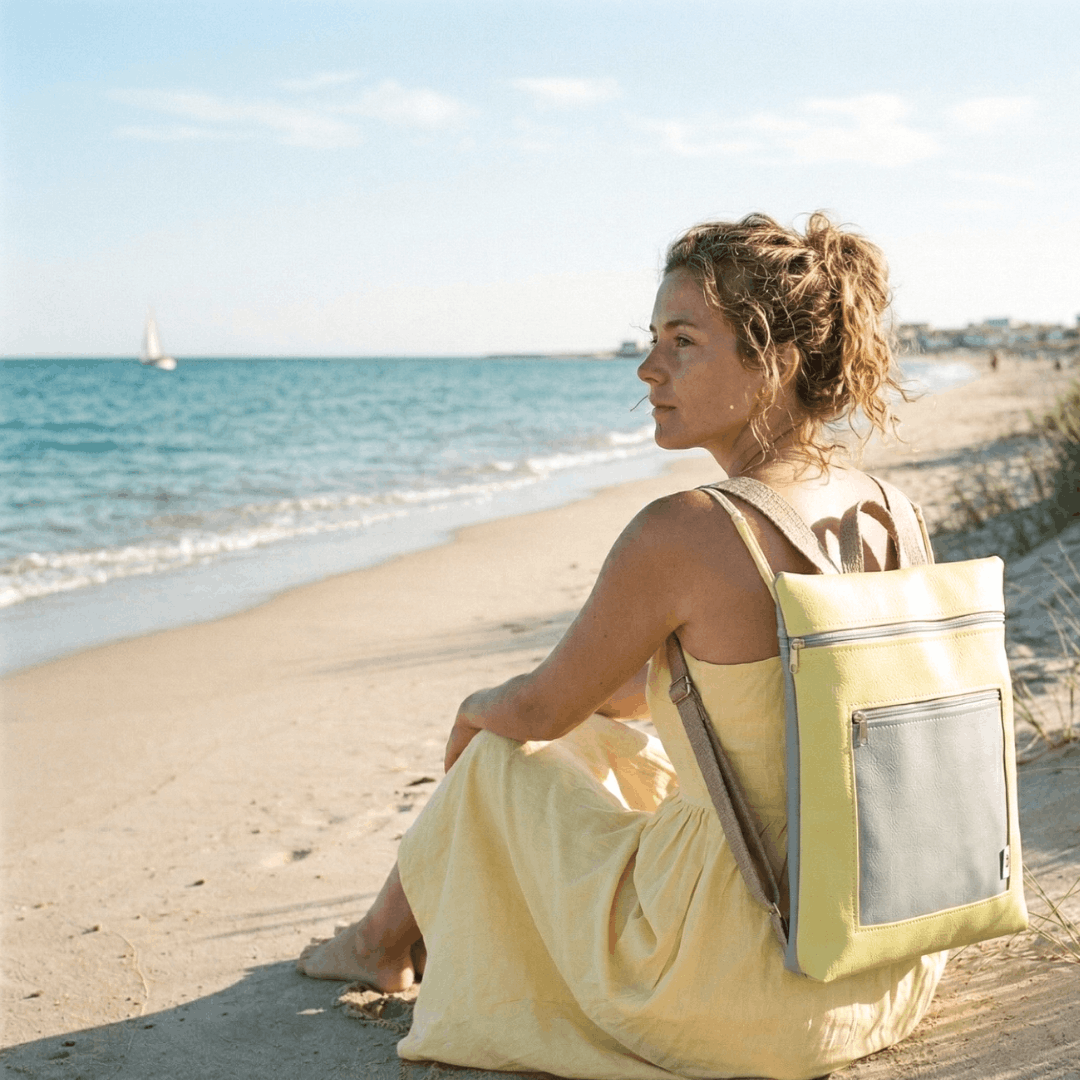 Mujer senatada en la playa mirando el mar y llevando una mochila artesanal de Cristina Alba